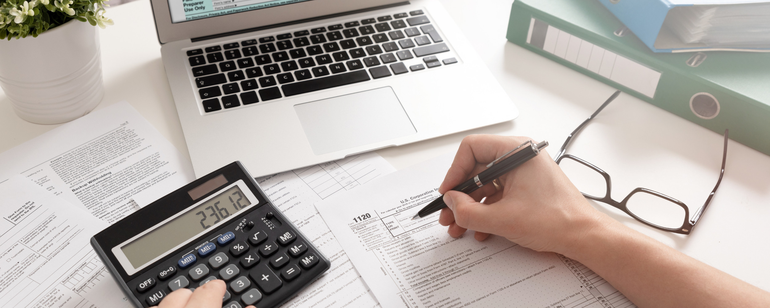 Picture of a calculator, open laptop and the hand of a person taking notes.