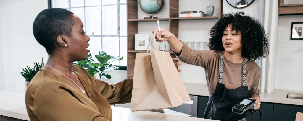 A woman hands a brown paper bag to another woman, after making a purchase at a retail store.