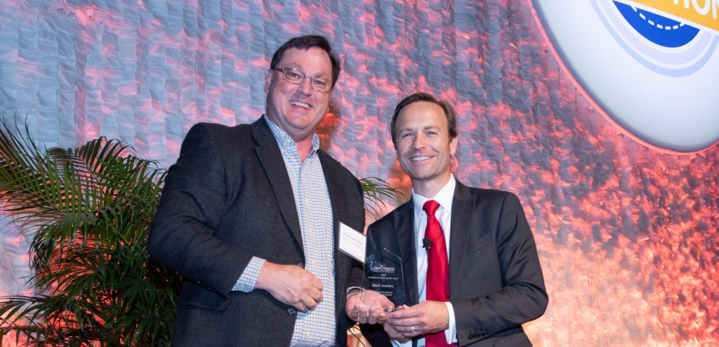 Two men standing, one of whom is holding an award.