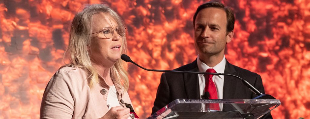 A woman stands at a microphone with a man behind her observing.