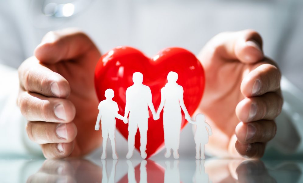 Paper cutouts of four people: two adults and two children, in front of a red heart with hands surrounding the heart