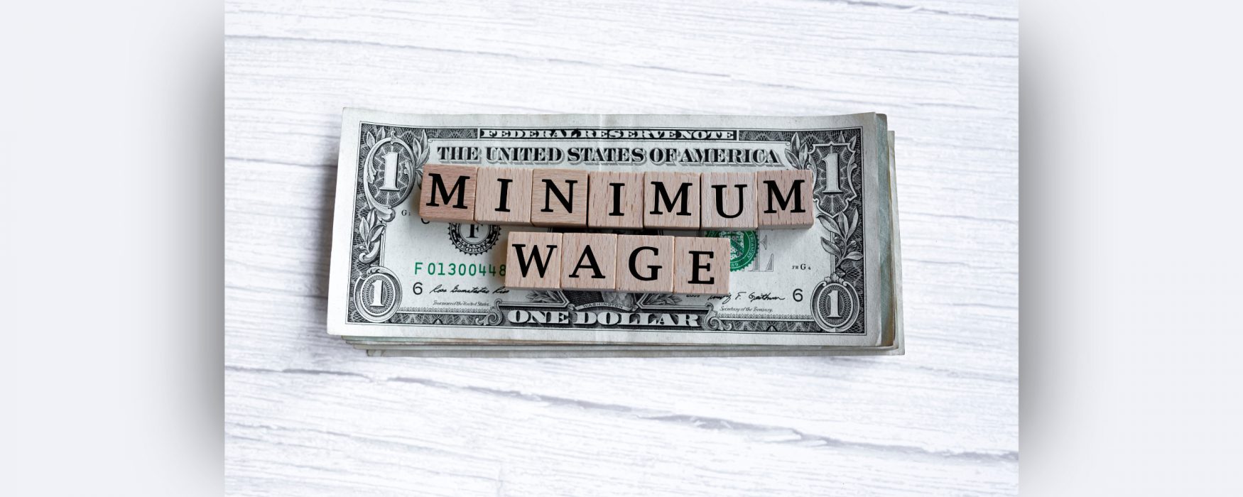 Image of a dollar bill with scrabble blocks on top of it, spelling out minimum wage