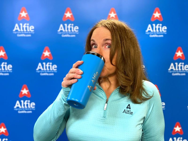 A woman named Bonnie Alfonso drinks from a logo branded cup in front of a step and repeat with the Alfie logo