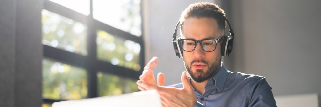 A man sits at a laptop with headphones on appearing to speak to someone on the screen